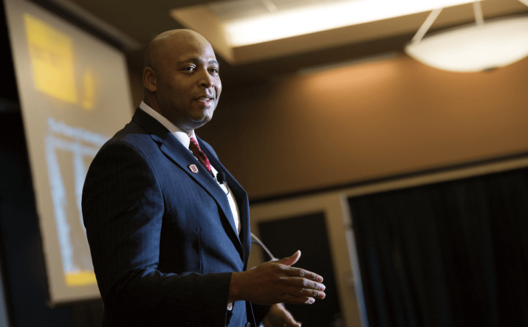 Man in blue suit with Bradley University pin delivering speech in front of screen